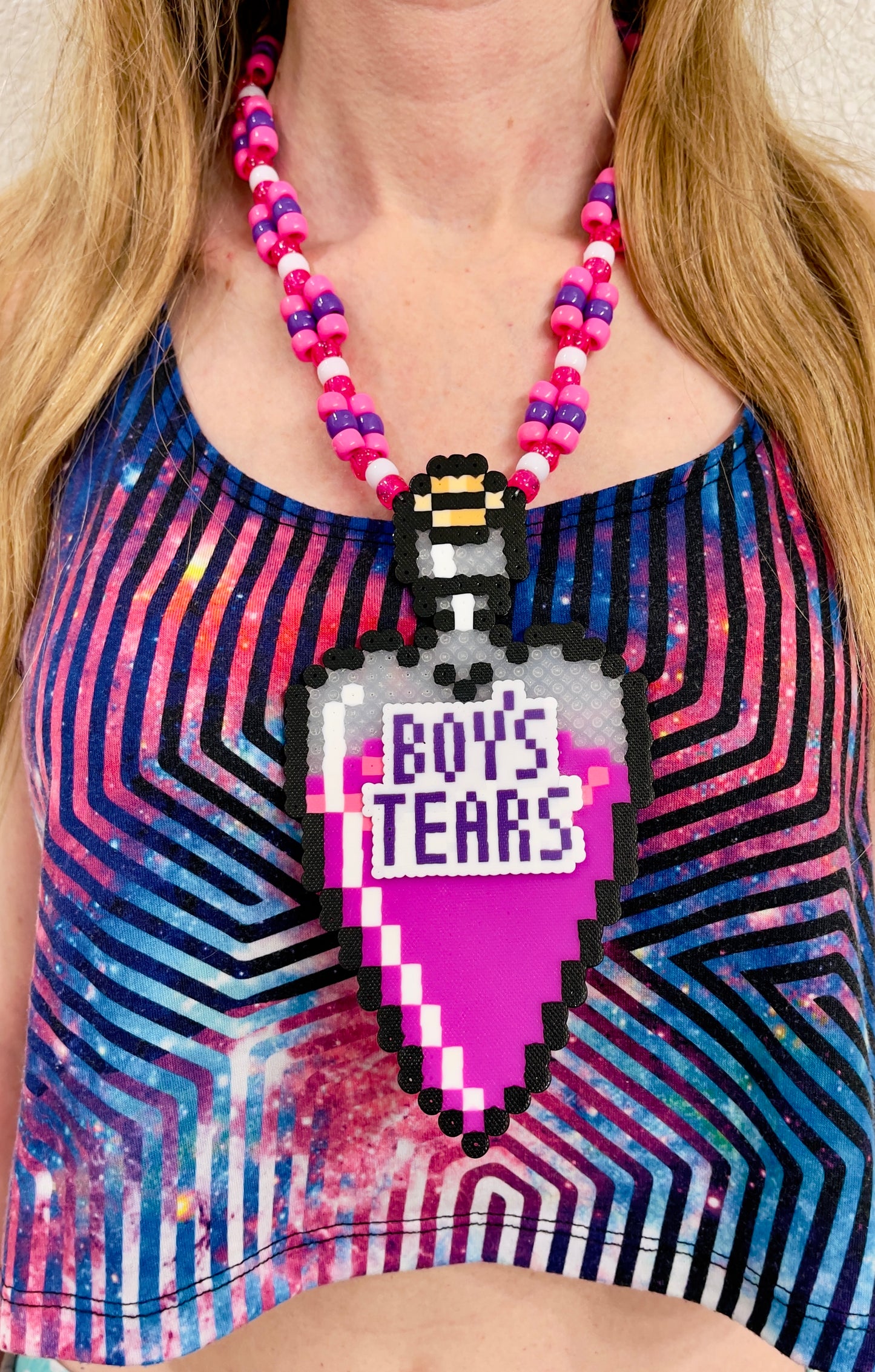 bottle in the shape of a heart that has a label on it that says Boys Tears made by Leaky Pixels. It is filled with a pink liquid. pink and purple perler necklace.