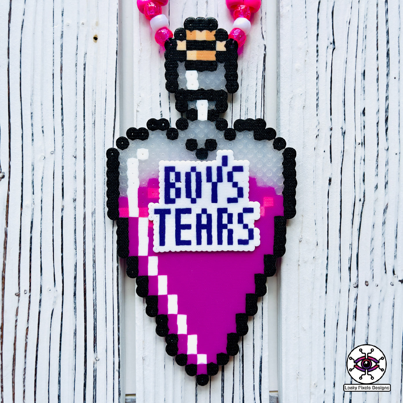 bottle in the shape of a heart that has a label on it that says Boys Tears made by Leaky Pixels. It is filled with a pink liquid. pink and purple perler necklace.