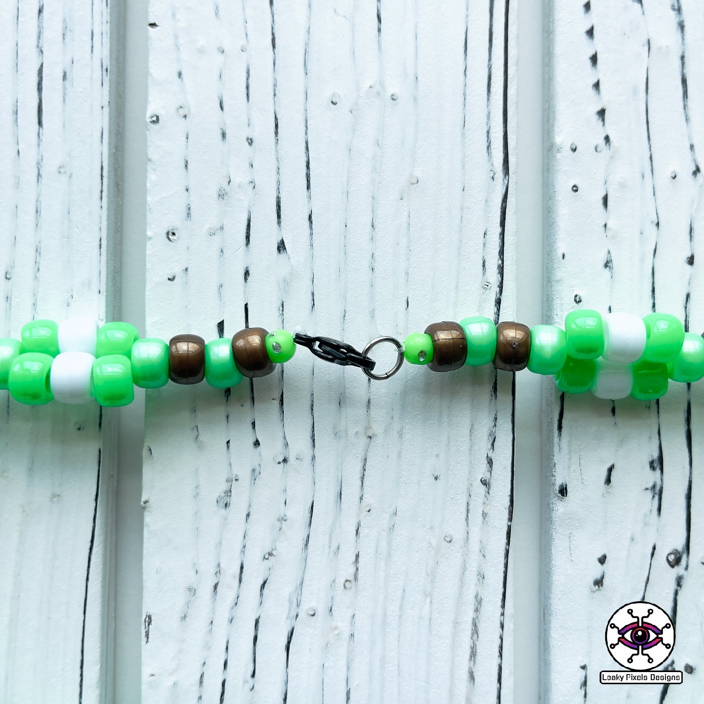 Baby grogu subtronics perler necklace. green yoda with cyclops eye and chaotic teeth. necklace is made with brown, white and green perler beads. He is wearing a brown outfit.
