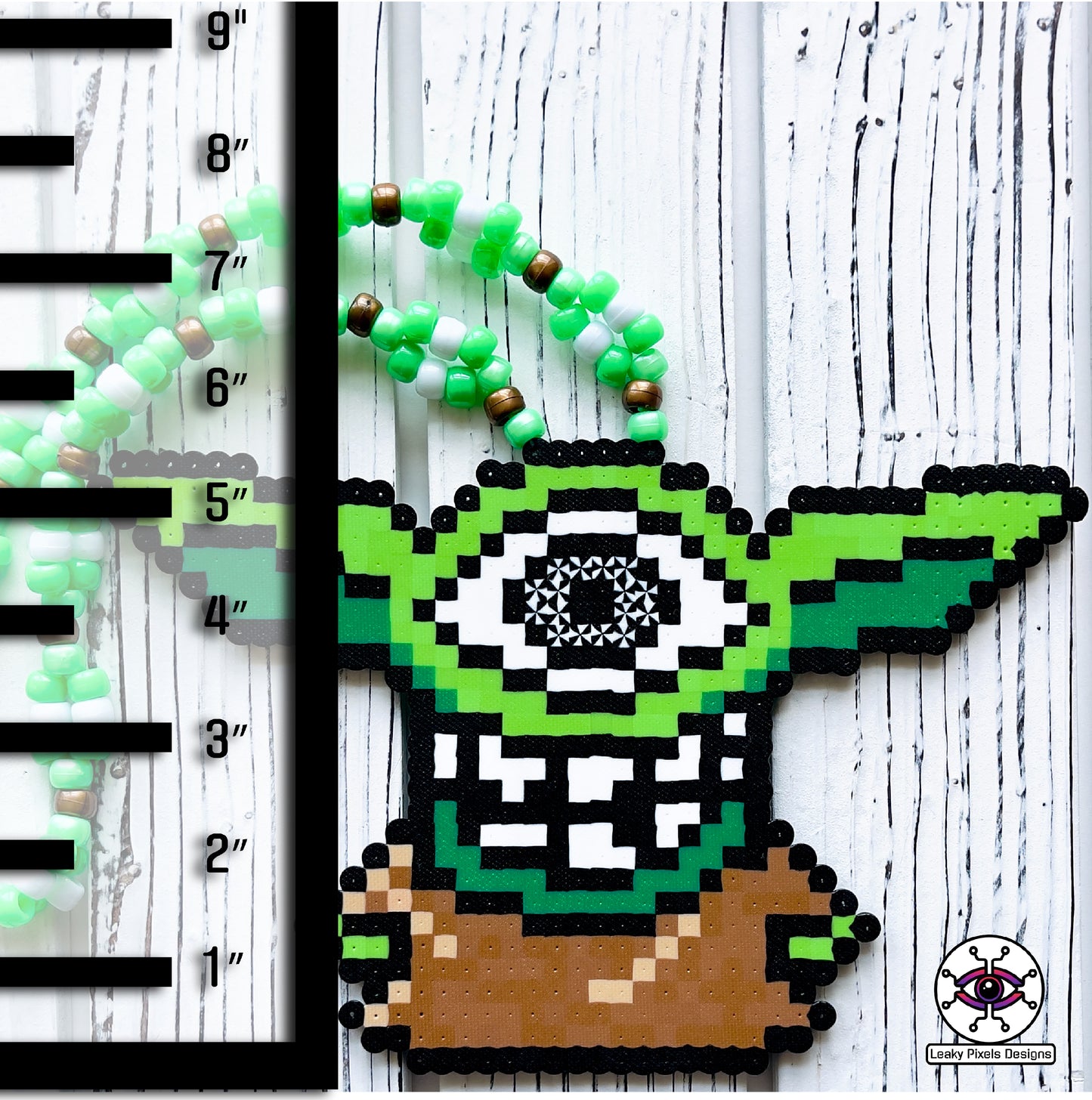 Baby grogu subtronics perler necklace. green yoda with cyclops eye and chaotic teeth. necklace is made with brown, white and green perler beads. He is wearing a brown outfit.