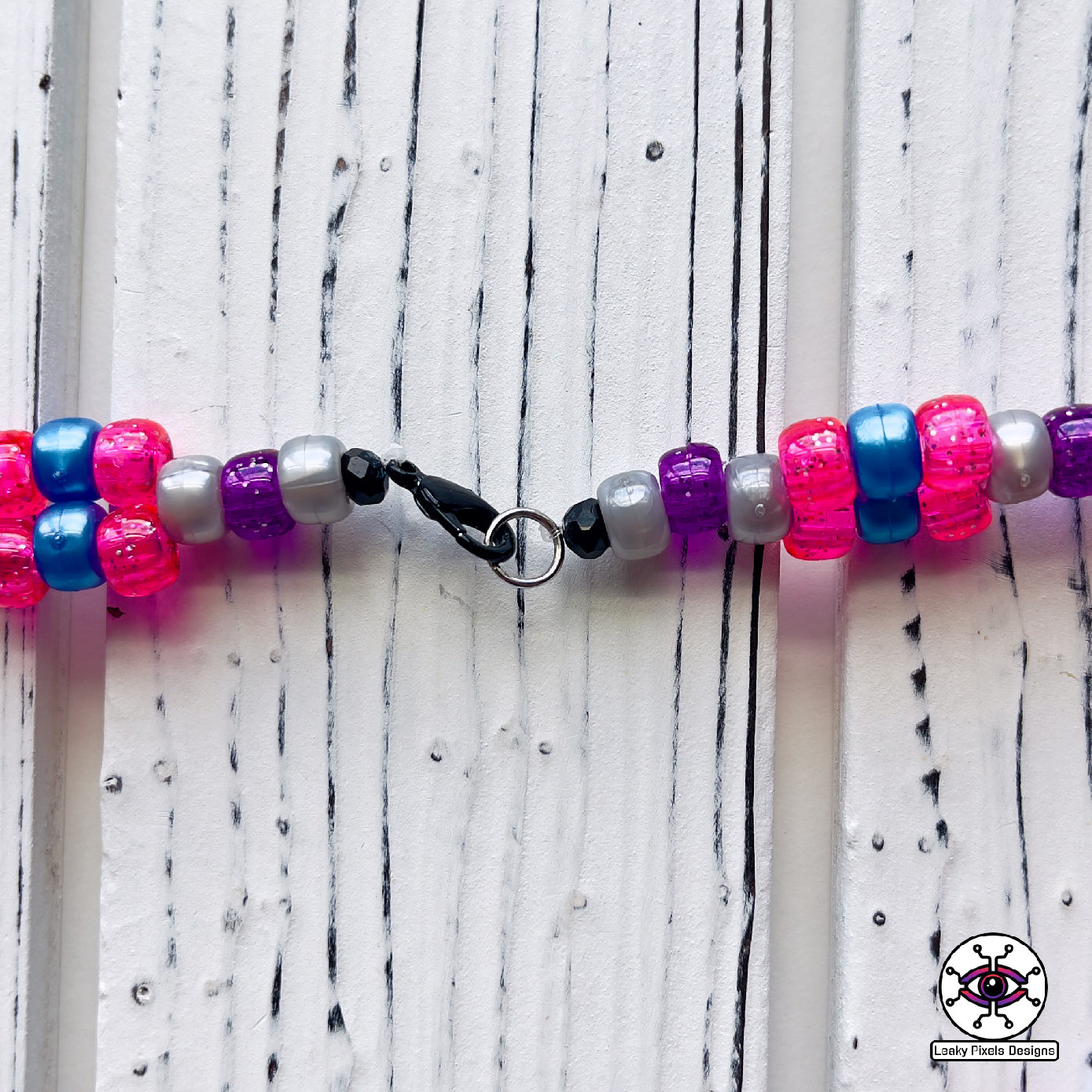 Seven Lions galaxy perler necklace. seven lions logo made of purple, pink, teal, blue and white starts. image of a galaxy. necklace is made of purple, blue, and pink pearl pony beads.