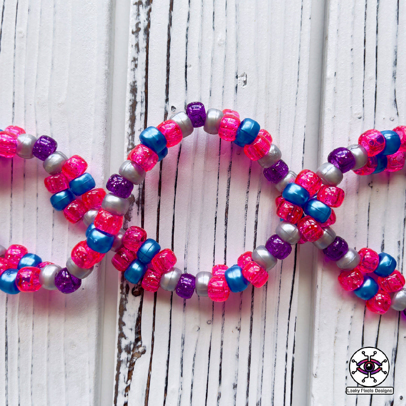 Seven Lions galaxy perler necklace. seven lions logo made of purple, pink, teal, blue and white starts. image of a galaxy. necklace is made of purple, blue, and pink pearl pony beads.