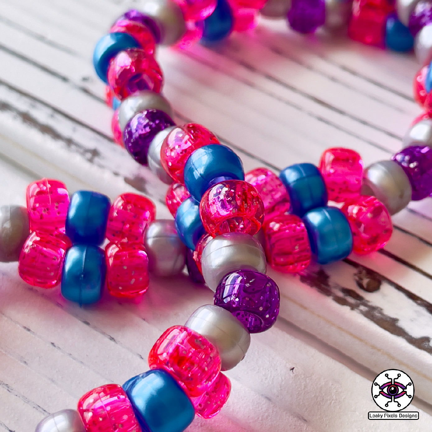 Seven Lions galaxy perler necklace. seven lions logo made of purple, pink, teal, blue and white starts. image of a galaxy. necklace is made of purple, blue, and pink pearl pony beads.