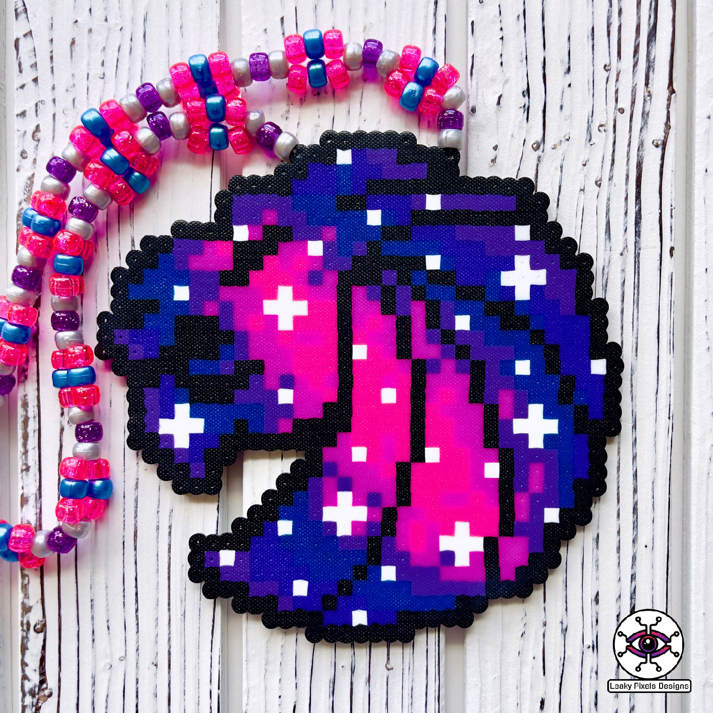 Seven Lions galaxy perler necklace. seven lions logo made of purple, pink, teal, blue and white starts. image of a galaxy. necklace is made of purple, blue, and pink pearl pony beads.