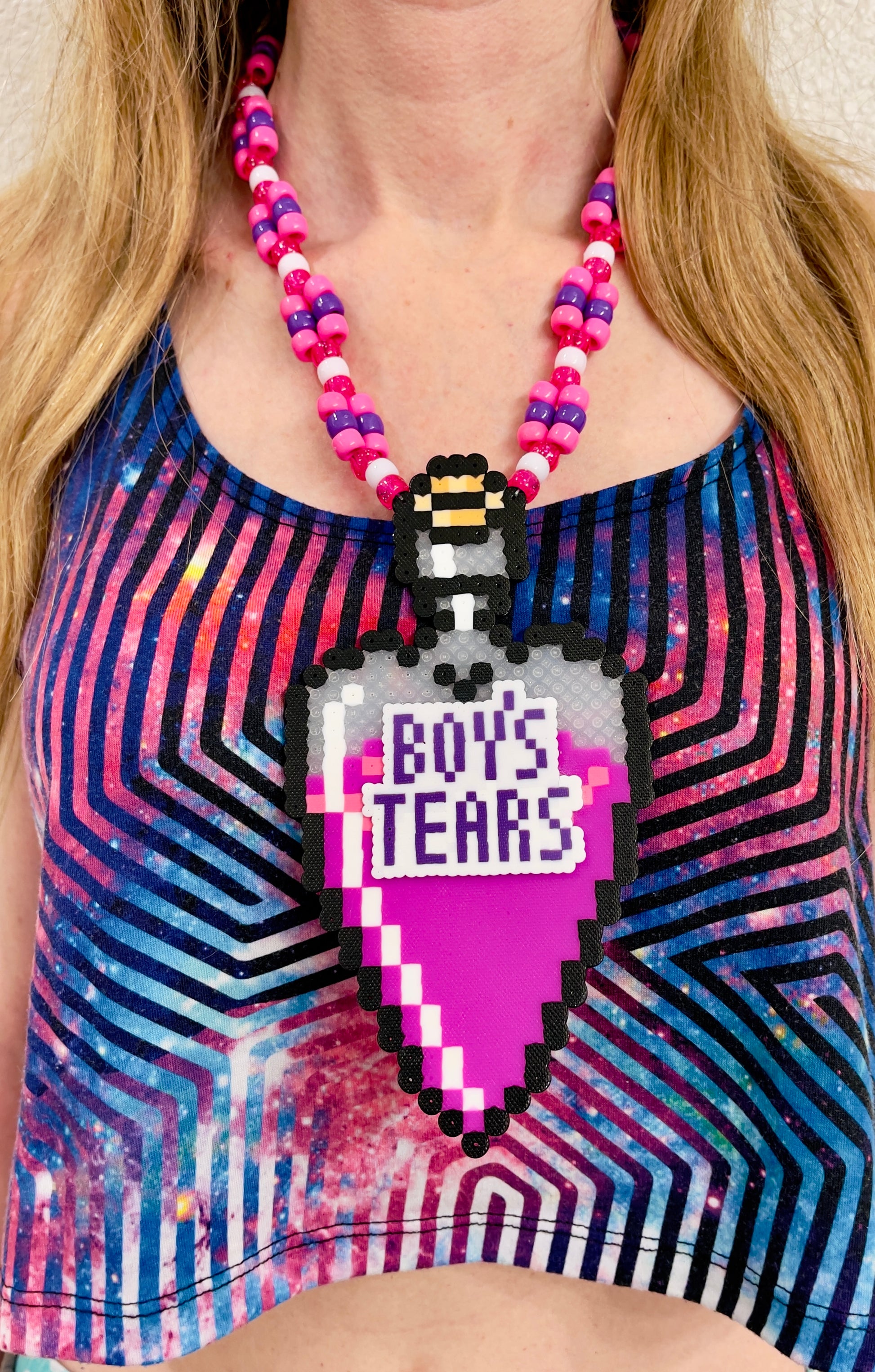 bottle in the shape of a heart that has a label on it that says Boys Tears made by Leaky Pixels. It is filled with a pink liquid. pink and purple perler necklace.