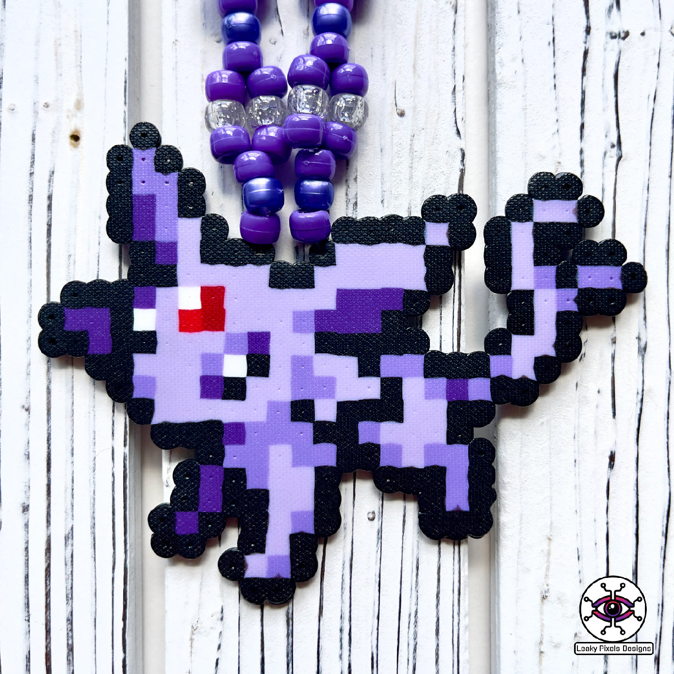 Espion perler necklace. Perler of the pokemon Espion that is cat like with fur colors of lavender and purple. Has a red gem stone on her forehead. Pony beads are purple for the necklace.