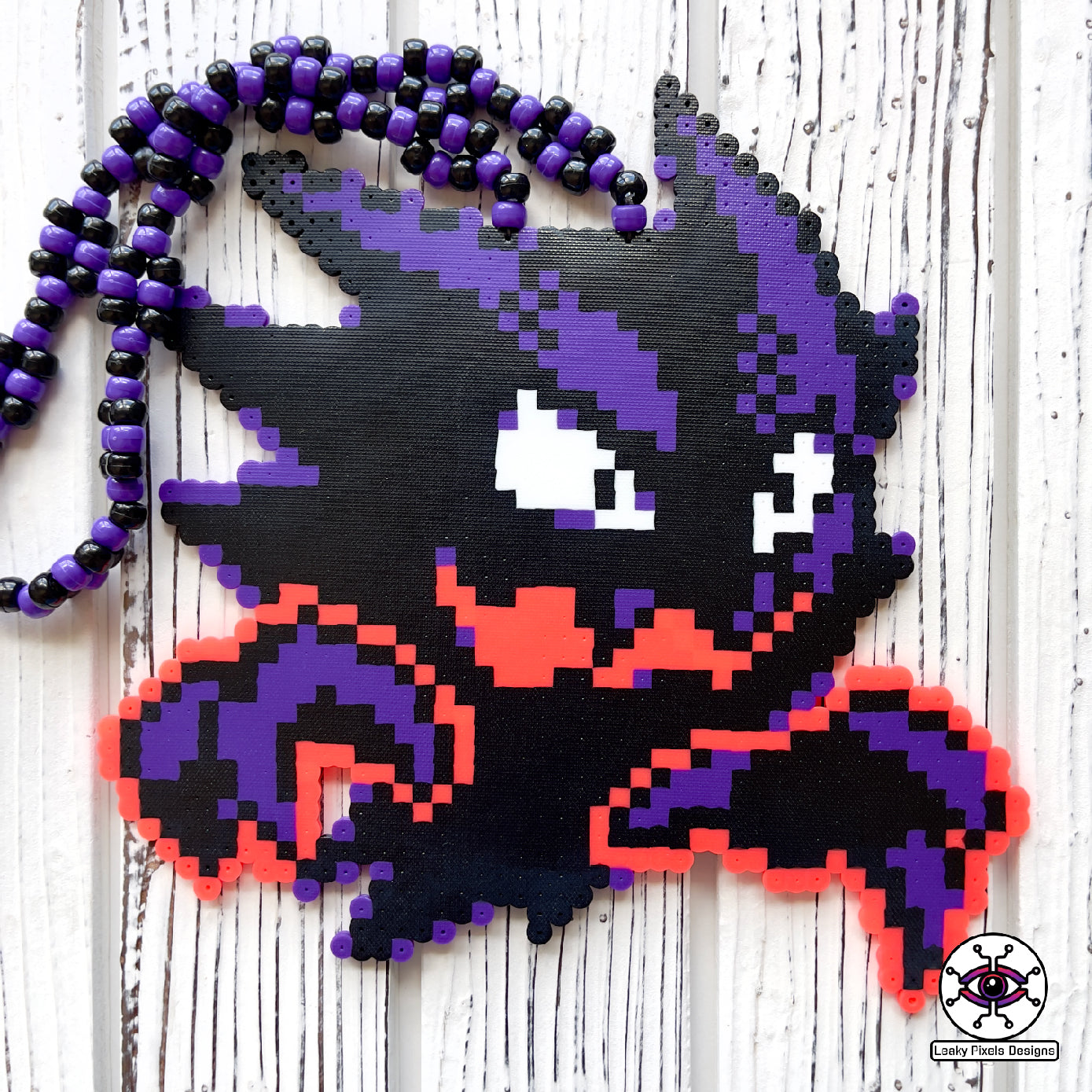 Haunter pokemon perler necklace. black and purple ghost with hot coral glowing hands. necklace is made of purple and black glitter pony beads on a nice wood background.